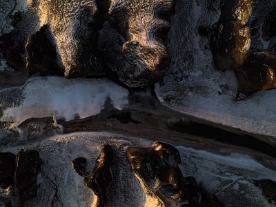 Aerial view of the dark rocks covered with snow and ice, illuminated by golden sunlight. The shadows cast on these rocks form unique patterns resembling animal heads. A small stream flows through it, adding to its natural beauty. Shot in high resolution using a Nikon D850 camera and an aperture setting of f/4, in the style of the photographer. --ar 4:3