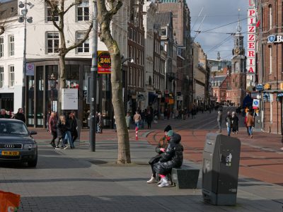 A photo shows the street in Amsterdam, with people walking around and sitting on benches next to trees. A black metal trash can is visible near an open pizzeria sign that says "PIbrightness" in red letters. In front there is a man wearing all dark who sits at one end while he looks out into traffic. On the other side are two women dressed like police officers standing guard outside their station as they watch over the road. One woman has her hands folded across her chest. --ar 4:3