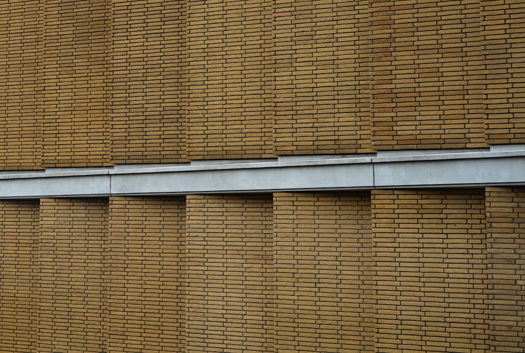 The exterior wall of the building is made up entirely of horizontal lines of brown bricks, with gray metal frames between them. The background has a simple and clean style, with no other elements in it. This brickwork creates an interesting texture on its surface. The style is reminiscent of modern architecture, depicted in real photography. –ar 64:43