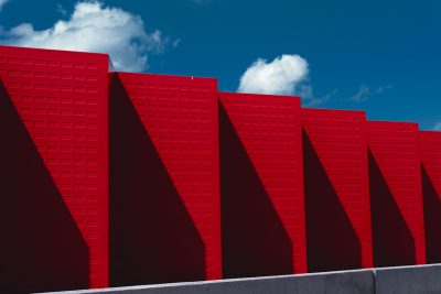 A photograph shows red wall panels with triangular shapes, arranged in an endless loop on the side of a building under a clear blue sky. The shadows cast by each panel create geometric patterns that add depth and texture to the composition. The photograph is in the style of [Ren Hang](https://goo.gl/search?artist%20Ren%20Hang). --ar 128:85