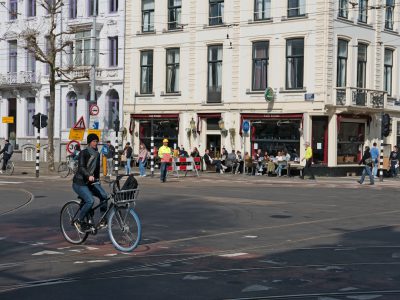 A Dutch city street with white buildings, cars and bicycles on the road, people sitting in outdoor cafes. A young man is riding his bike across an intersection while wearing jeans and black leather jacket. The woman next to him has blue wheels wrapped around her bicycle's front wheel for protection from dirt. In background there are pedestrians walking down both sides of each sidewalk and some traffic signs indicating speed America, China --ar 4:3