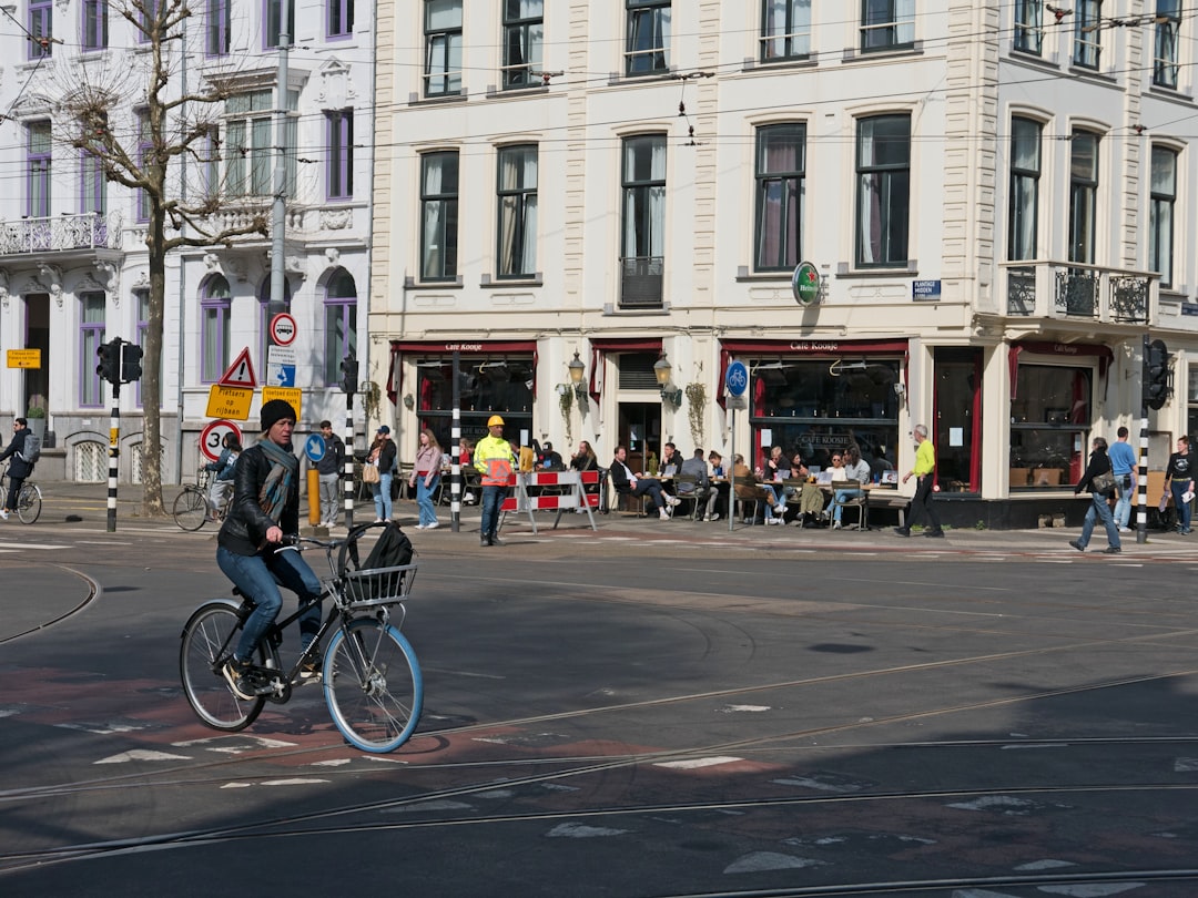 A Dutch city street with white buildings, cars and bicycles on the road, people sitting in outdoor cafes. A young man is riding his bike across an intersection while wearing jeans and black leather jacket. The woman next to him has blue wheels wrapped around her bicycle’s front wheel for protection from dirt. In background there are pedestrians walking down both sides of each sidewalk and some traffic signs indicating speed America, China –ar 4:3