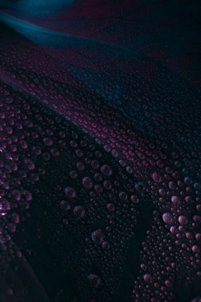 Dark purple background, water droplets on the car hood, with subtle pink and blue light reflections, creating an abstract pattern. The surface of the texture is covered in tiny bubbles that reflect in various colors under different angles. It creates a mysterious atmosphere. Closeup view of macro photography in the style of abstract patterns. --ar 85:128