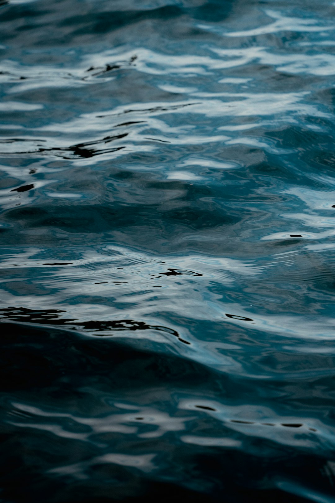 A closeup of the dark blue water surface, with ripples and reflections in it. The background is blurred and dark, creating an atmosphere full of mystery and tranquility. Soft lighting illuminates the calm waves on the sea level, adding depth to the scene. High definition photography style, highlighting details and textures.,,in –ar 85:128