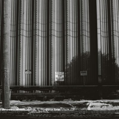 Black and white photograph of the side wall of an industrial building with vertical stripes. Next to it is a sign reading "ITCHJumpParkatWishlyInferior" and another saying "Paige global". In front there are some train tracks. The photograph was shot on a Leica M6 with 35mm film, displaying grain at high resolution in a hyperrealistic style.