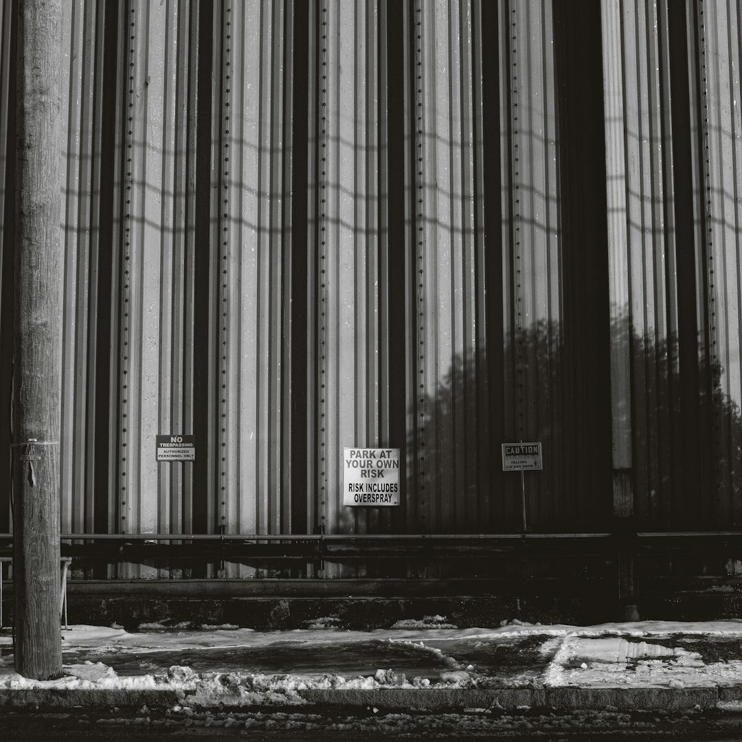 Black and white photograph of the side wall of an industrial building with vertical stripes. Next to it is a sign reading “ITCHJumpParkatWishlyInferior” and another saying “Paige global”. In front there are some train tracks. The photograph was shot on a Leica M6 with 35mm film, displaying grain at high resolution in a hyperrealistic style.