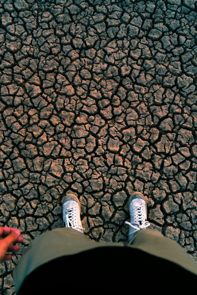 In the middle of an apocalyptic desert, there is cracked earth and dry rivers on both sides. A person wearing white sneakers stands in front with their feet facing up to show shoes from above. The perspective angle shows that they stand directly under the sky without any trees or plants around them. They have short hair and wear green pants, while holding hands for warmth. The image has a cinematic style, using a wide angle lens with natural light and a low saturation tone to create a sad atmosphere, in the style of a wideangle lens, natural light, low saturation tone, sad atmosphere. --ar 85:128