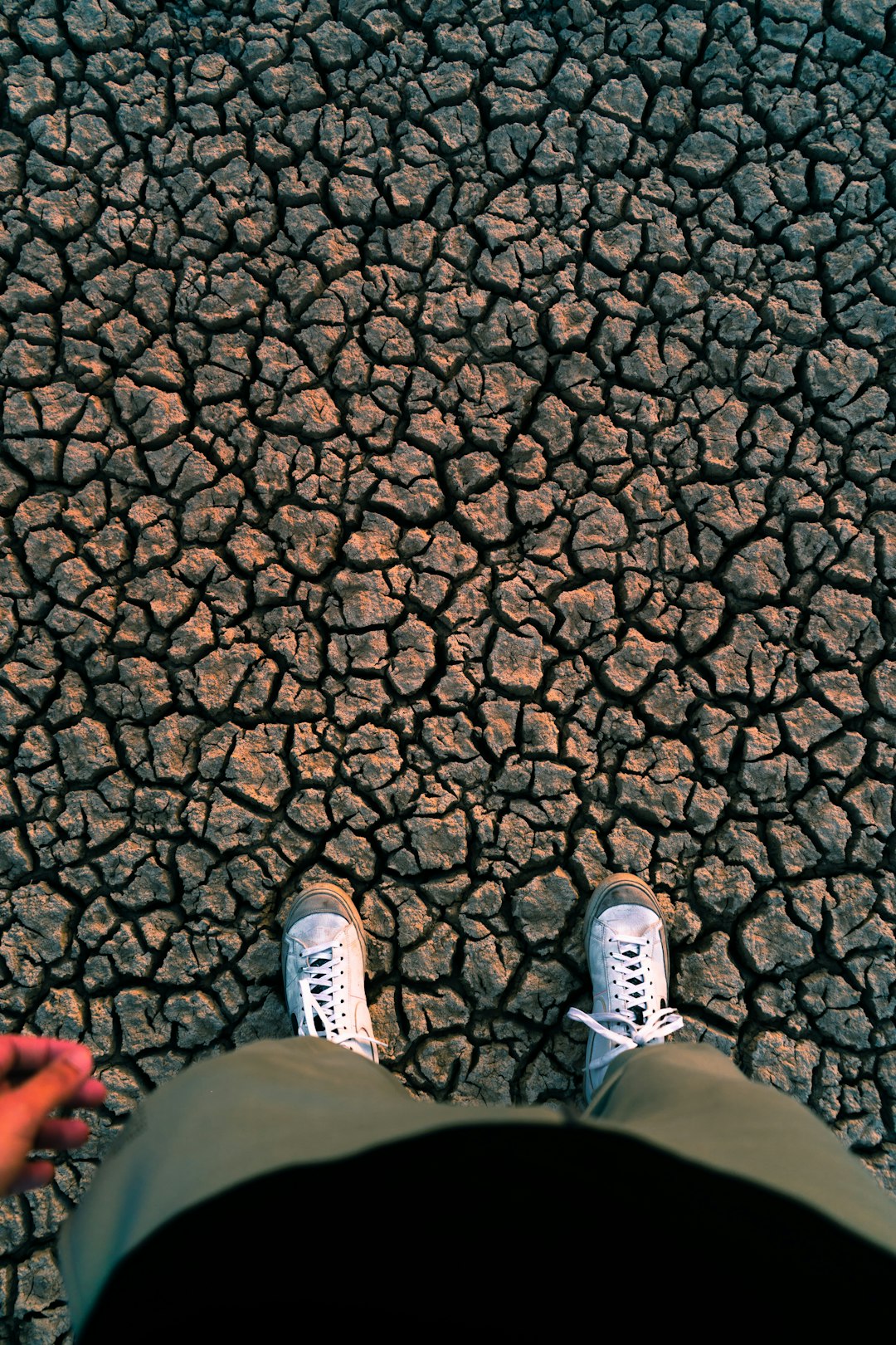 In the middle of an apocalyptic desert, there is cracked earth and dry rivers on both sides. A person wearing white sneakers stands in front with their feet facing up to show shoes from above. The perspective angle shows that they stand directly under the sky without any trees or plants around them. They have short hair and wear green pants, while holding hands for warmth. The image has a cinematic style, using a wide angle lens with natural light and a low saturation tone to create a sad atmosphere, in the style of a wideangle lens, natural light, low saturation tone, sad atmosphere. –ar 85:128