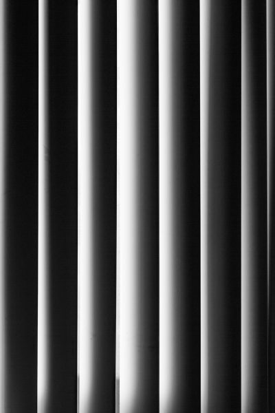 A black and white photograph of vertical blinds, with the shadows cast by sunlight creating an abstract pattern on their surface. The focus is sharp against a solid background, highlighting the texture and design details of each blind. This composition creates a minimalist aesthetic that emphasizes the beauty in simplicity in the style of minimalism. --ar 85:128