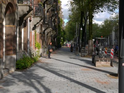 A street in Amsterdam with cobblestone, old buildings and trees on the right side. Construction work is going on in one part of the sidewalk. A woman is walking holding a shopping bag at the far end of the road. A motorcycle is parked next to an iron fence on the left hand side of the road. It is a sunny day. The photo was taken from the front. --ar 4:3