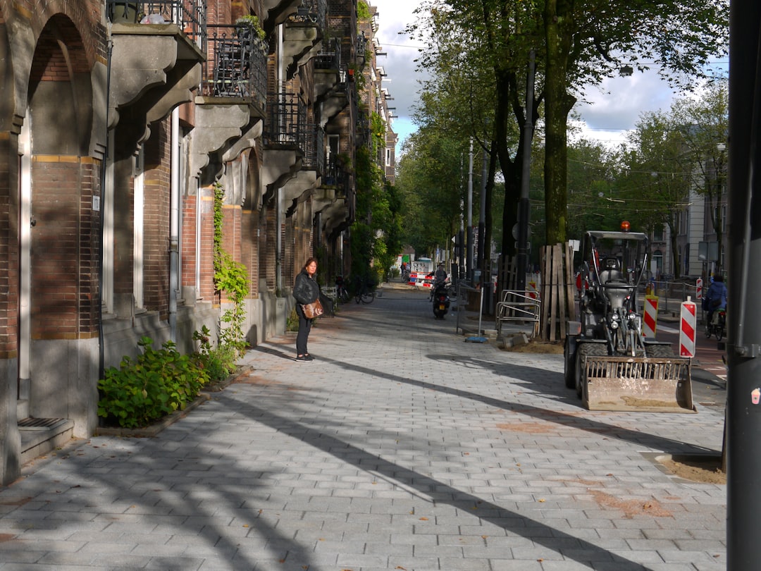 A street in Amsterdam with cobblestone, old buildings and trees on the right side. Construction work is going on in one part of the sidewalk. A woman is walking holding a shopping bag at the far end of the road. A motorcycle is parked next to an iron fence on the left hand side of the road. It is a sunny day. The photo was taken from the front. –ar 4:3