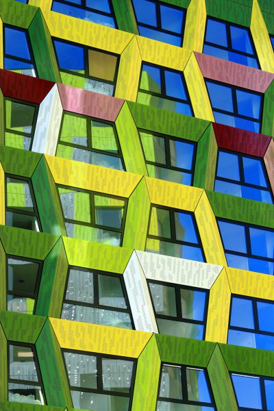 In the heart of Amsterdam, in front of an office building made entirely from colorful hexagons with white frames, vibrant green and yellow hues contrast against the blue sky. The facade is adorned with geometric patterns that create a visually striking effect, showcasing modern architecture at its best. This unique architectural masterpiece captures attention while providing breathtaking views for all who gaze upon it. The focus is on the facade. --ar 85:128