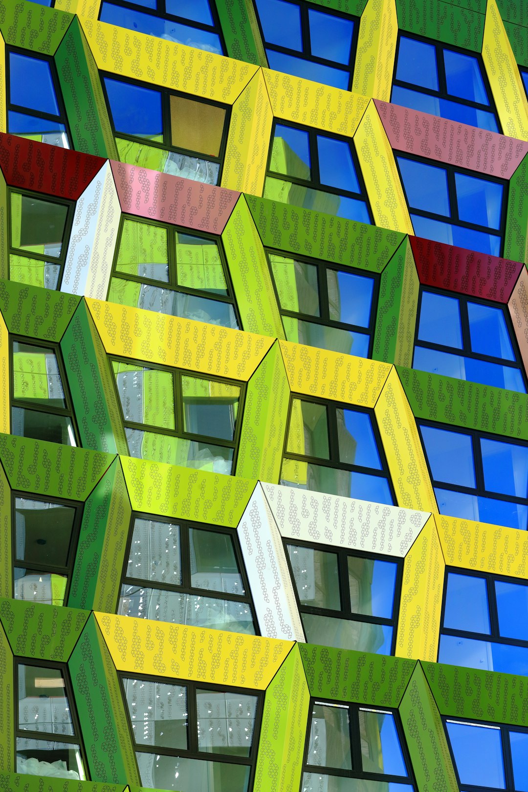 In the heart of Amsterdam, in front of an office building made entirely from colorful hexagons with white frames, vibrant green and yellow hues contrast against the blue sky. The facade is adorned with geometric patterns that create a visually striking effect, showcasing modern architecture at its best. This unique architectural masterpiece captures attention while providing breathtaking views for all who gaze upon it. The focus is on the facade. –ar 85:128