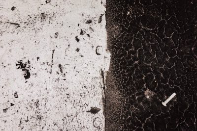 A closeup of the texture on an asphalt surface, with white and black details, featuring small cracks in between and a single piece of cigarette merchant at one side. The background is plain, emphasizing the contrast between concrete and paint. This image has been created using the aesthetic style reminiscent of vintage photography, with a focus on realism and texture detail. It's perfect for adding depth to various design projects that need to convey urban or industrial vibes. --ar 128:85