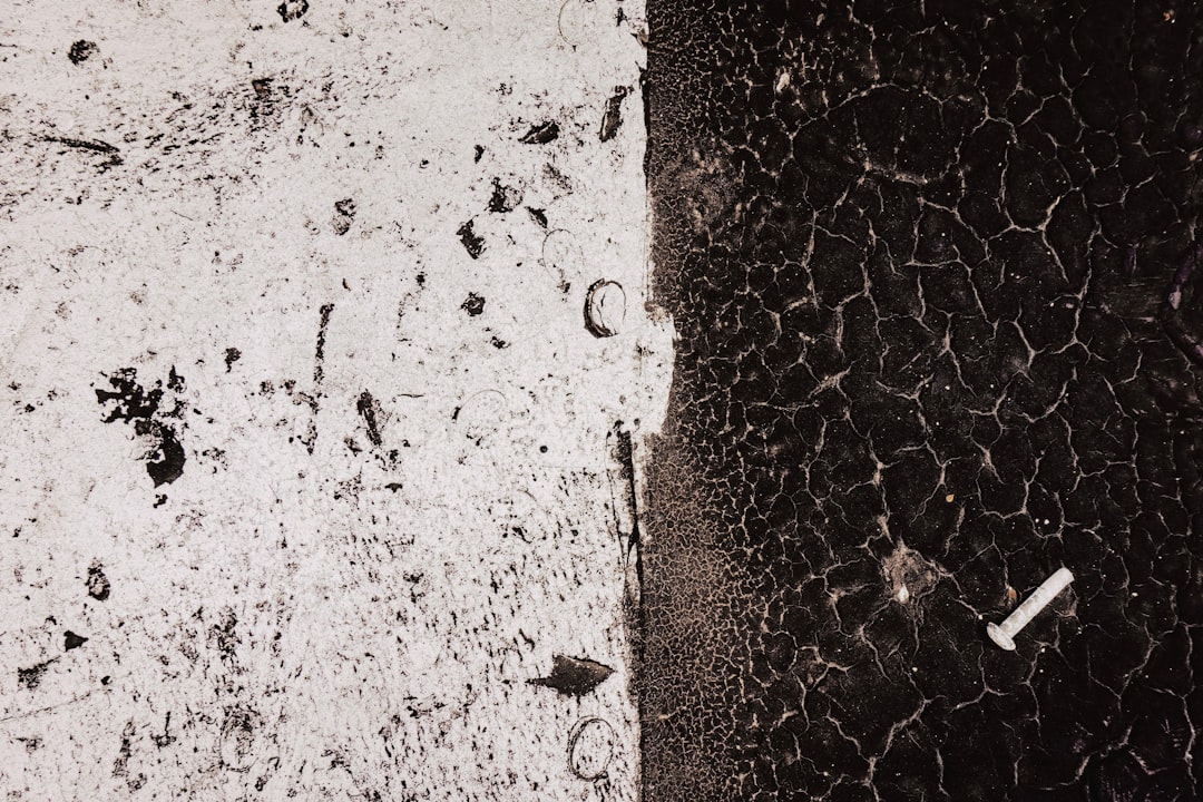 A closeup of the texture on an asphalt surface, with white and black details, featuring small cracks in between and a single piece of cigarette merchant at one side. The background is plain, emphasizing the contrast between concrete and paint. This image has been created using the aesthetic style reminiscent of vintage photography, with a focus on realism and texture detail. It’s perfect for adding depth to various design projects that need to convey urban or industrial vibes. –ar 128:85