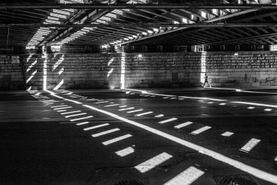 A black and white photograph of an empty urban space under the bridge, with sunlight casting dramatic shadows on its floor, creating geometric patterns that add depth to the scene. The composition is symmetrical, focusing attention on one side where there's no person visible but instead just lines and shapes. This creates a contrast between lightness and darkness in a city setting. --ar 128:85