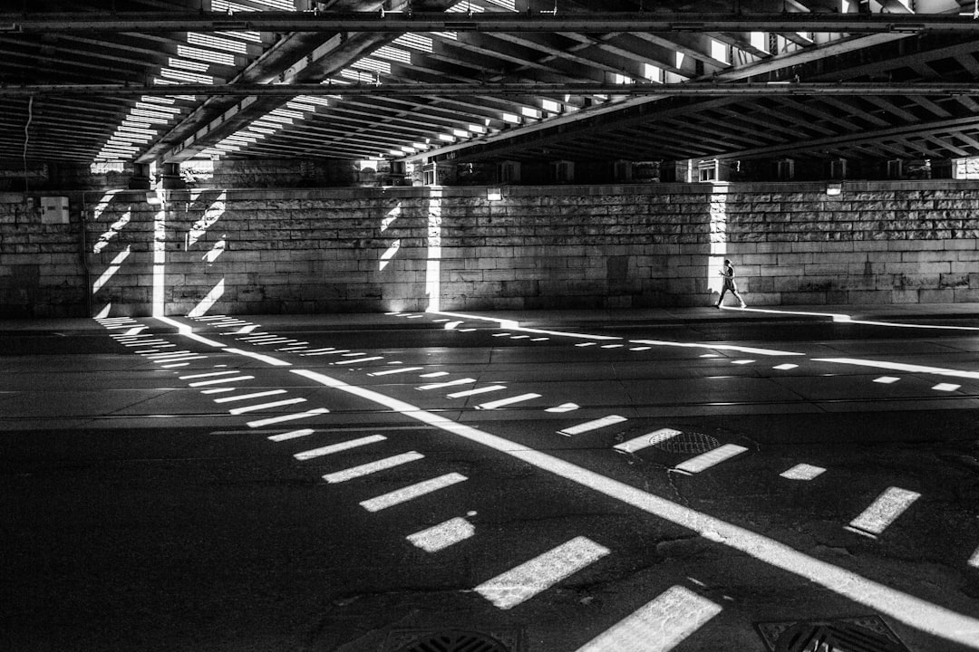 A black and white photograph of an empty urban space under the bridge, with sunlight casting dramatic shadows on its floor, creating geometric patterns that add depth to the scene. The composition is symmetrical, focusing attention on one side where there’s no person visible but instead just lines and shapes. This creates a contrast between lightness and darkness in a city setting. –ar 128:85