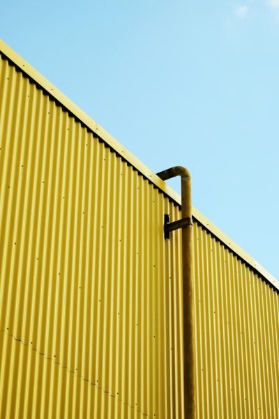 A photo of a yellow corrugated metal wall with one black pipe against a clear blue sky, taken from a low angle, in the style of minimalism and an industrial style. --ar 85:128