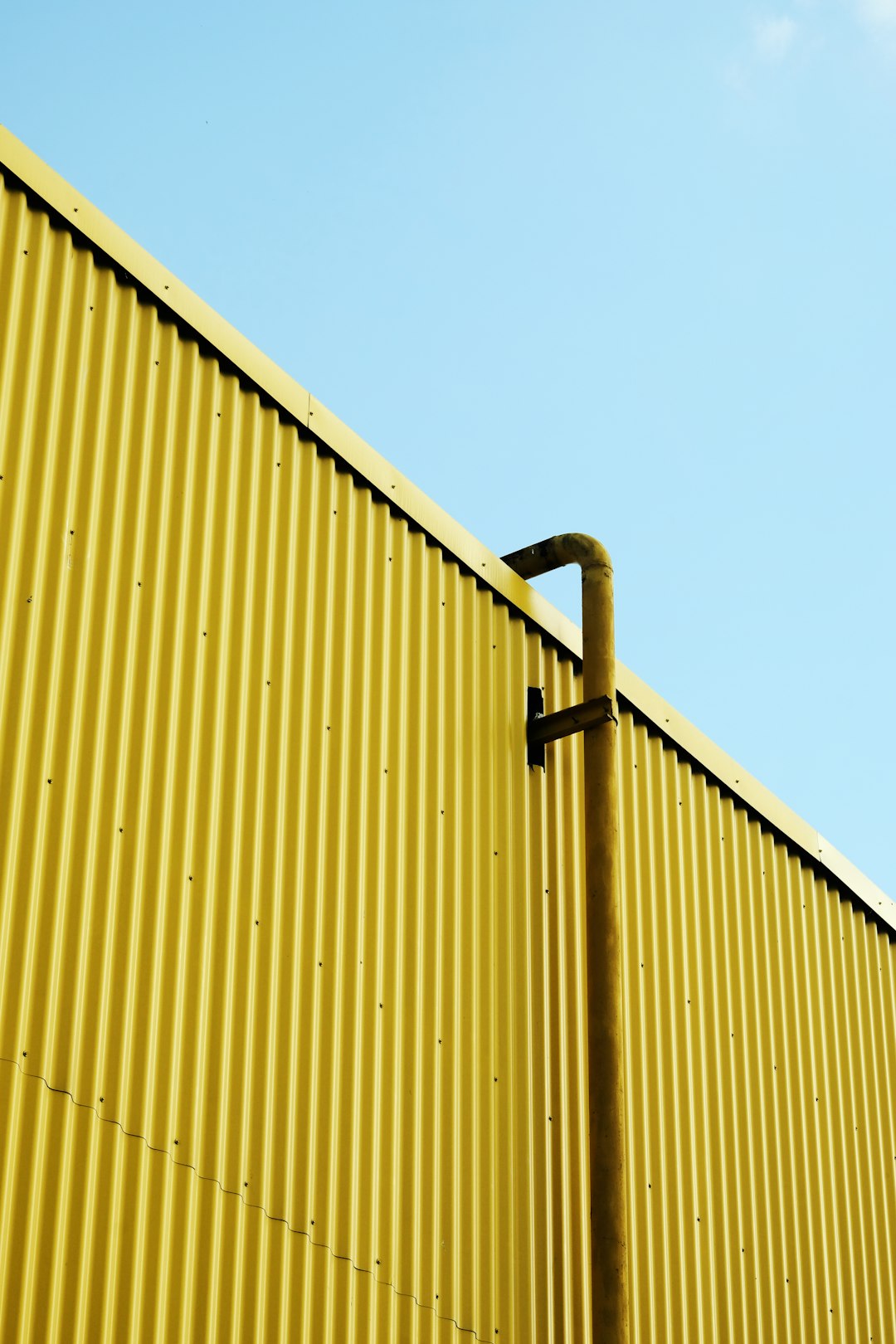 A photo of a yellow corrugated metal wall with one black pipe against a clear blue sky, taken from a low angle, in the style of minimalism and an industrial style. –ar 85:128