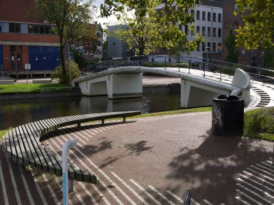 A small white bridge over the river in Amsterdam, on one side of it is an open space with some benches and green grass. On the other side there's a black trash can. In front of the entrance to the park there’s a straight striped red tile floor. There’s also water flowing under arches of the concrete bridge. It looks like a sunny day, buildings visible behind the trees. The photo was taken from eye level in the style of an Impressionist painter. --ar 4:3