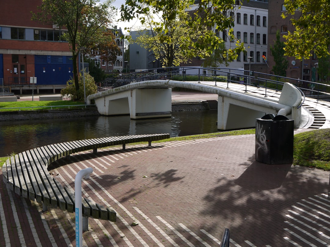 A small white bridge over the river in Amsterdam, on one side of it is an open space with some benches and green grass. On the other side there’s a black trash can. In front of the entrance to the park there’s a straight striped red tile floor. There’s also water flowing under arches of the concrete bridge. It looks like a sunny day, buildings visible behind the trees. The photo was taken from eye level in the style of an Impressionist painter. –ar 4:3