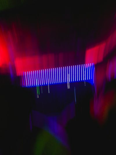 A long exposure photo shows an ultraviolet light bar shining in the dark, with people dancing underneath it. There is motion blur on their bodies and hands. The light creates blue lines that make waves or patterns on the black background. A hint of red color comes from one corner of the room. --ar 95:128