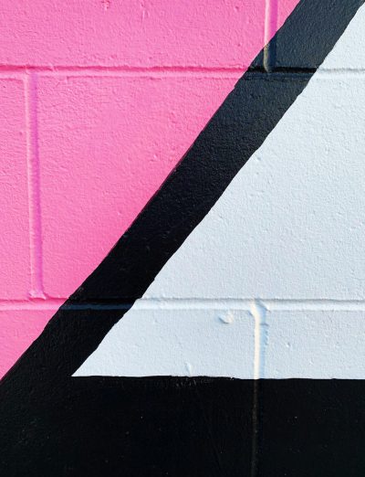 A closeup of the corner of an outdoor wall painted in pink, black and white graffiti. The design is minimalist with bold lines and geometric shapes, creating sharp angles that contrast against each other to create visual depth. A diagonal line from one color block at the left side to another color block on the right side creates movement within the composition in the style of minimalist graffiti artists. --ar 49:64