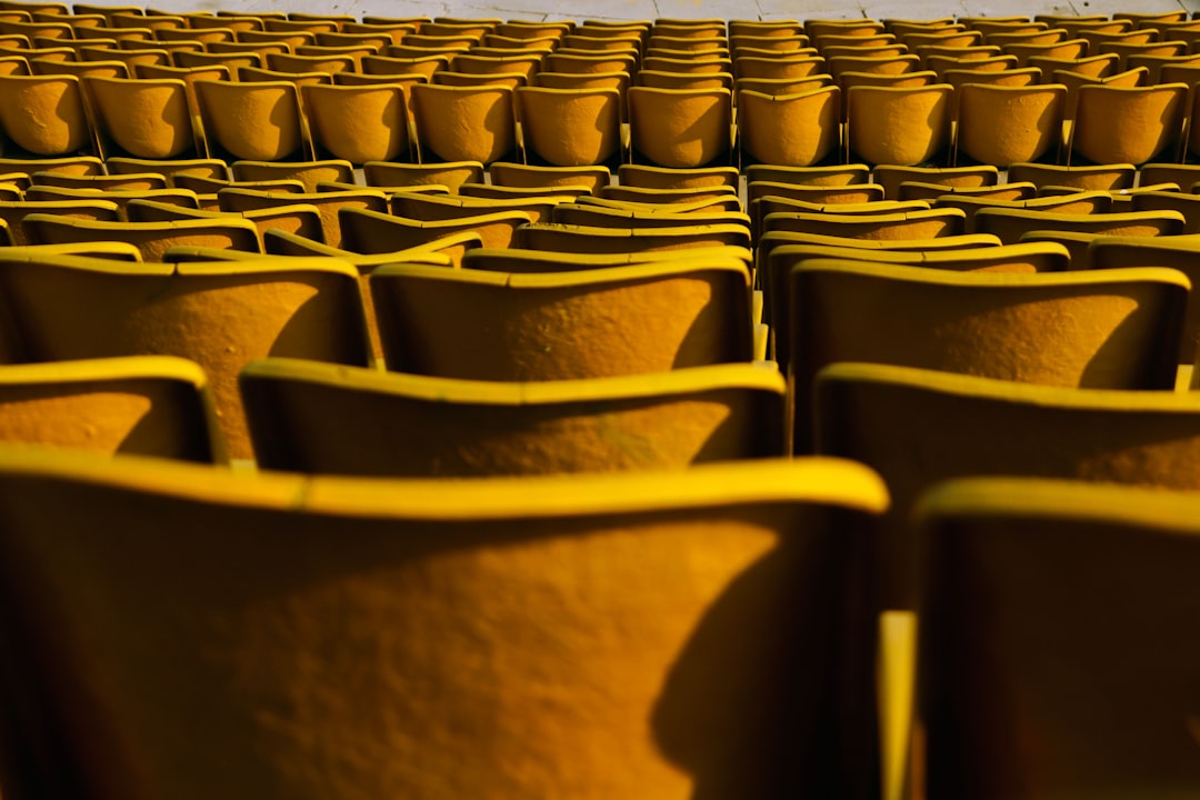 A photograph of yellow seats in an outdoor stadium, taken from the perspective of someone sitting on one chair looking at another row of chairs during golden hour with warm sunlight casting long shadows and highlighting each seat’s texture and shape. The background is blurred to focus attention on the individual seating arrangement. Taken with a Canon EOS5D Mark III camera with macro lens, capturing intricate details of textures and colors in the style of Canon. –ar 128:85