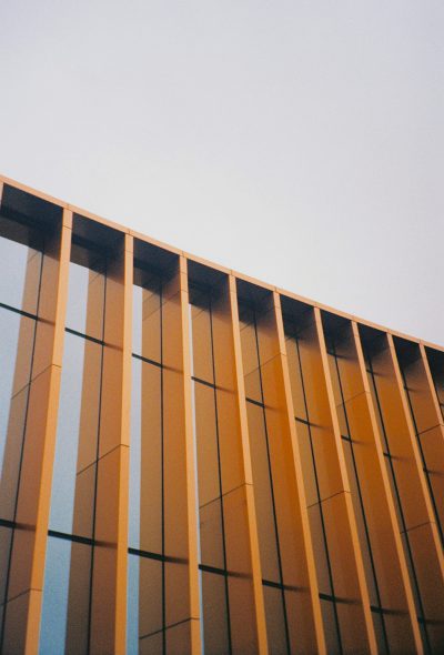 A photo of the facade of an office building made from vertical slats in metal and wood, with light orange color. The sky is clear and blue, shot on Kodak Gold 400 film --ar 43:64