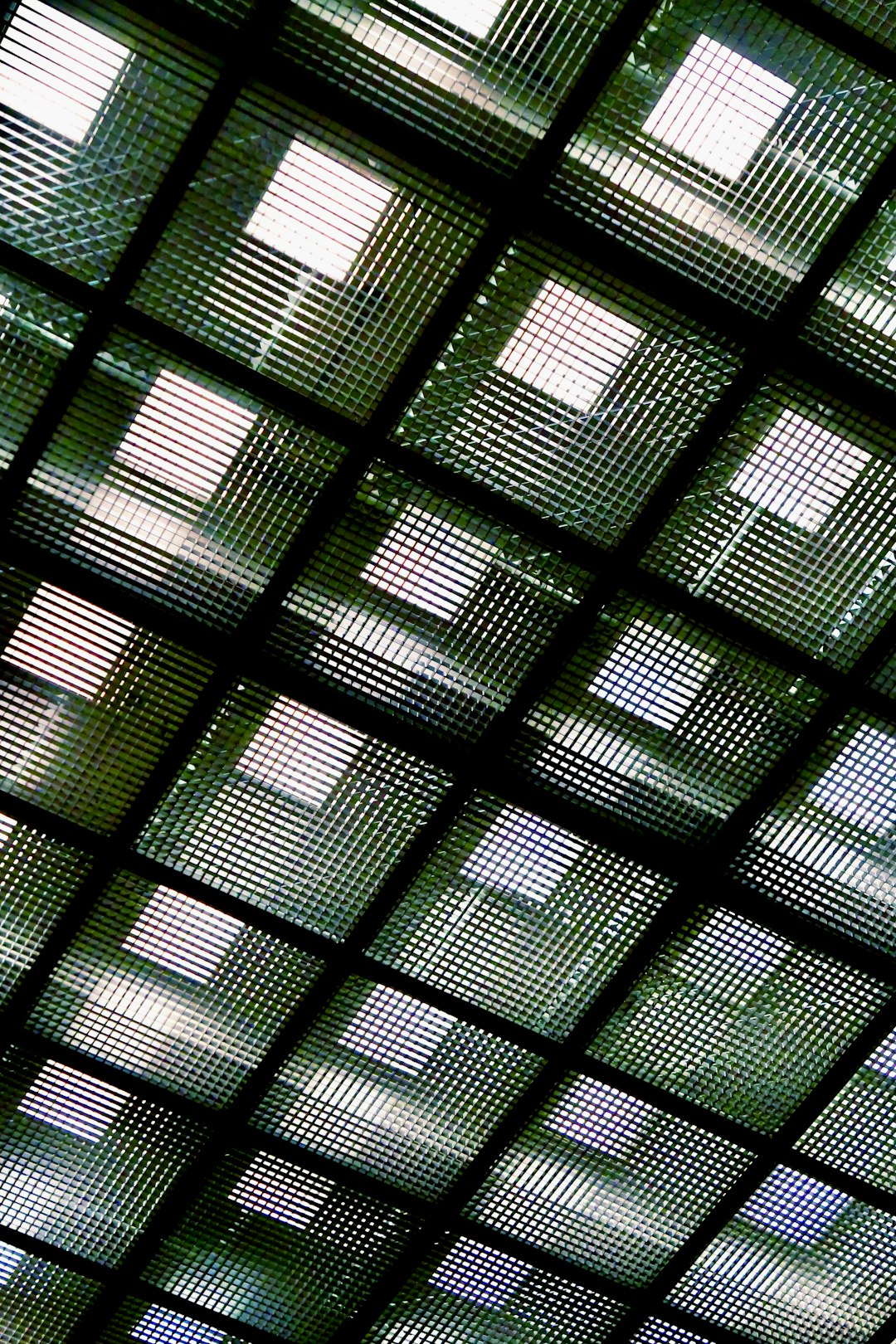 A photograph of the ceiling made from glass blocks, the glass is green and has grid patterns. The patterned light shines through the glass, creating shadows on the walls below. In sharp focus, the texture of each block is shown in detail. Taken with an iPhone camera, the image captures the intricate details of the glass blocks against the dark background, in the style of an iPhone camera. –ar 85:128