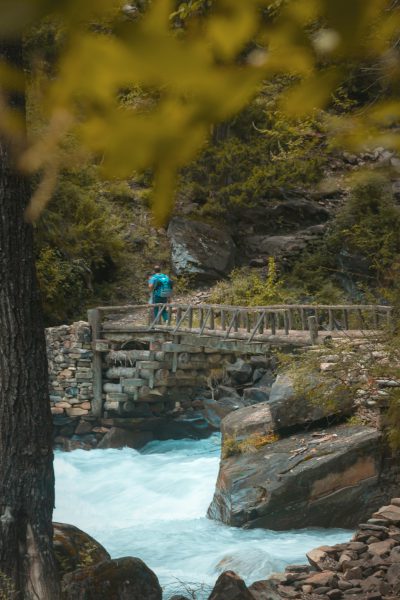 A hiker crossing an old wooden bridge over the fast flowing white water of And liebe's River in Patagonia, Chile. The scene is captured from behind with vibrant green trees and rocks on either side of the river, and there’s an attractive woman wearing blue shorts standing at one end of the bridge looking down into the river as she takes off her backpack to get ready for hiking. In the style of cinematic photography, beautiful, high resolution, sharp focus. --ar 85:128