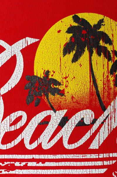 A red t-shirt with the word "Beach" in white, with palm trees and a sunset behind it. The design is distressed and textured to give an old school feel. It's in a retro style that evokes nostalgia from vintage beachwear advertising posters. This print would be perfect for summer events or vacation gear, in the style of retro beachwear advertising posters. --ar 21:32