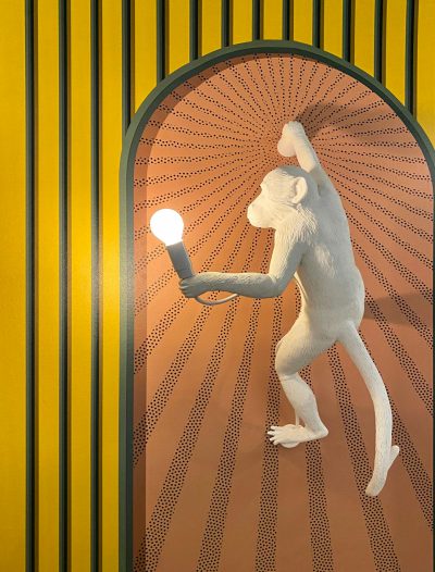 A white monkey holding an arched lamp is hanging on the wall, which has yellow and orange stripes with dots. The background features geometric shapes and lines. It was designed in the style of fashion designer Seusso HD, with high resolution and detailed details. Aerial photography was used to capture its full body, showing it in various poses. In front of him stands another light bulb, which adds visual contrast. This photo will be suitable for use as advertising material or interior decoration. --ar 97:128