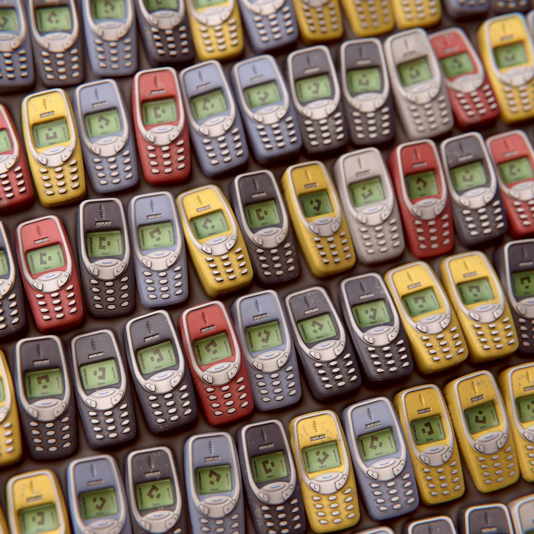 A pattern of small, colorful and worn nokias from the early ’90s with their display showing number”5″, arranged in rows on top of each other. The background is dark gray.