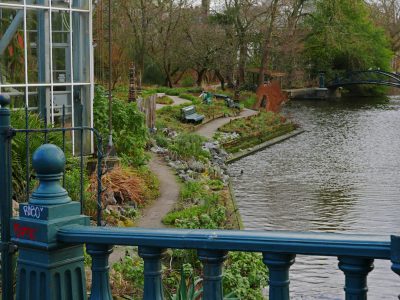 A view of the canal from an ornate blue and green bridge, overlooking an urban community garden with raised beds for vegetables and herbs along one side, surrounded by trees and some birds in flight, in winter with sparse leaves or no foliage on both sides of the path leading to it. The photo is taken at eye level looking down onto the waterway, taken with a wide-angle lens on a Canon EOS camera in the style of a realistic painter. --ar 4:3