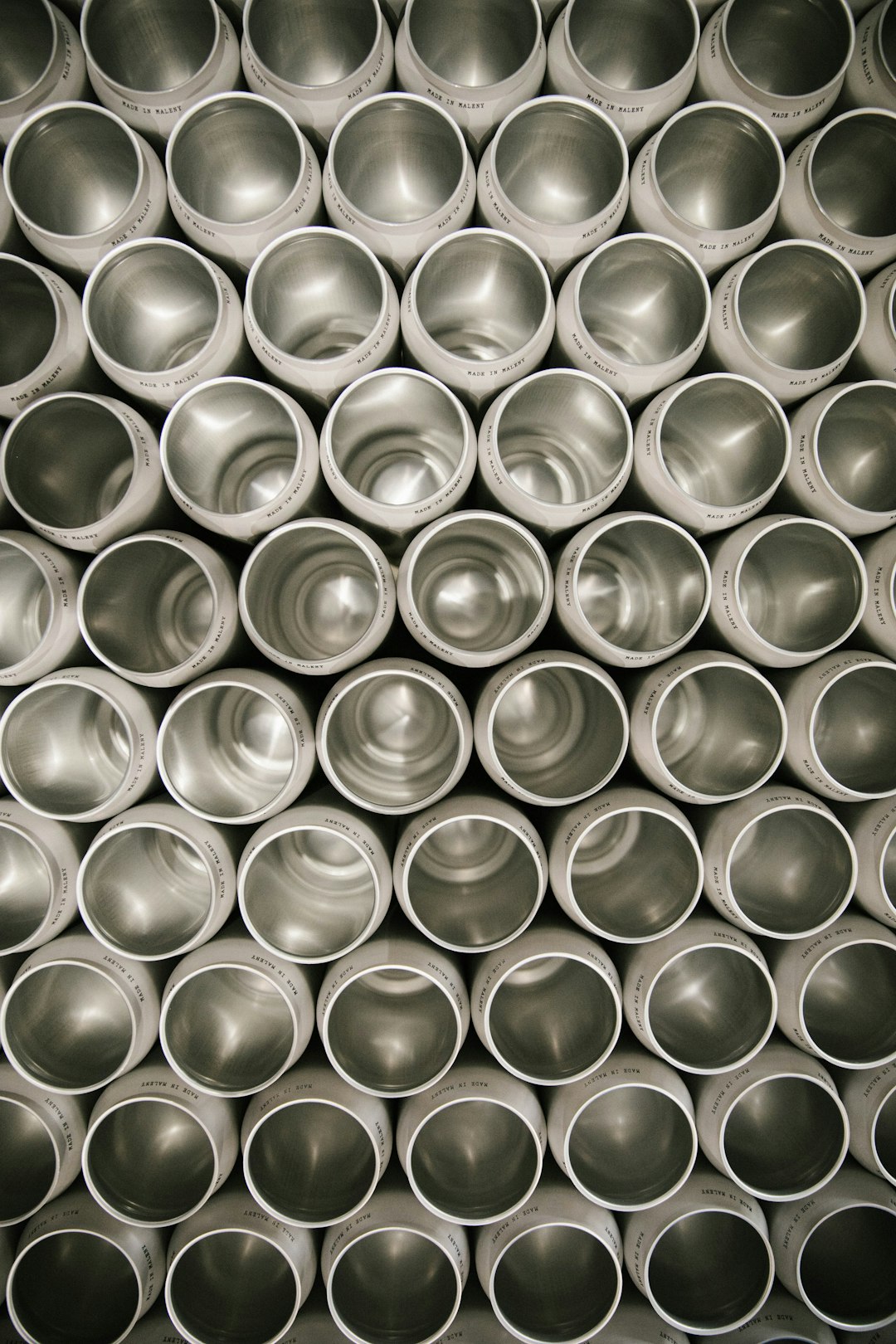 A symmetrical pattern of metal cups stacked on top of each other, arranged in neat rows and creating an abstract background. The metallic texture is visible through the open mouthed tubes, adding depth to the composition. –ar 85:128