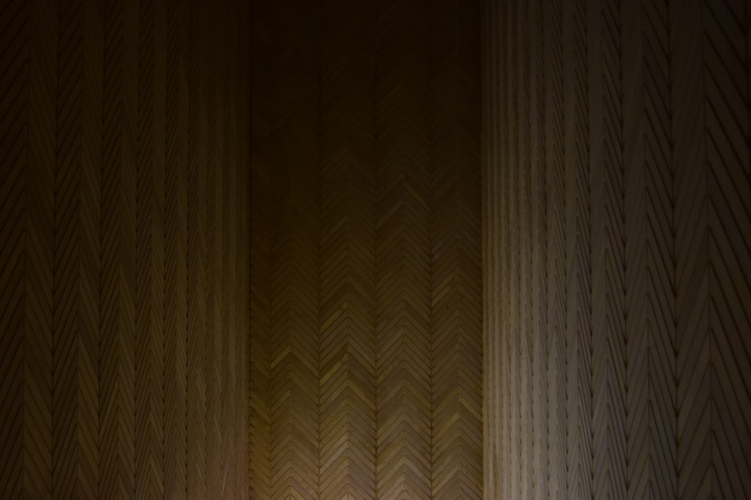 A wall with a herringbone pattern, illuminated by soft lighting. The pattern is made of wood and has dark tones. It’s a closeup shot that captures the texture and detail of each panel in the wall. This scene evokes an atmosphere of warmth and comfort. The lighting casts gentle shadows on the wooden surface, adding depth to the composition. In front of it stands a person wearing casual attire, looking at their phone screen in the style of casual attire, looking at their phone screen. –ar 128:85