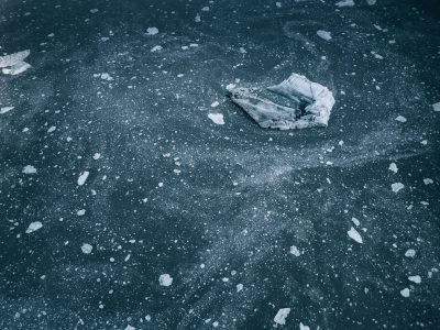 Flat ice on a lake, with small floating pieces of broken glass and debris visible. A top view with a dark blue, monochrome style and low contrast and saturation, photographed in the style of a photorealistic image shot with a Sony Alpha A7 III camera. --ar 128:95