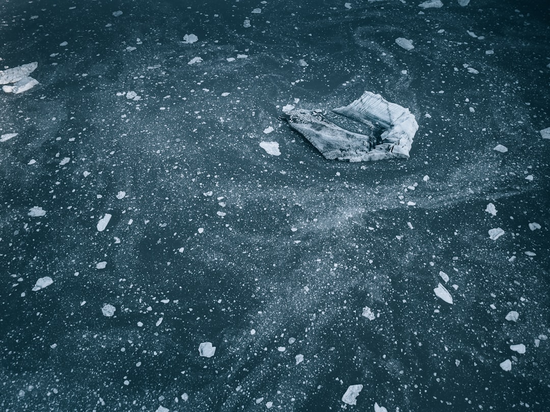 Flat ice on a lake, with small floating pieces of broken glass and debris visible. A top view with a dark blue, monochrome style and low contrast and saturation, photographed in the style of a photorealistic image shot with a Sony Alpha A7 III camera. –ar 128:95