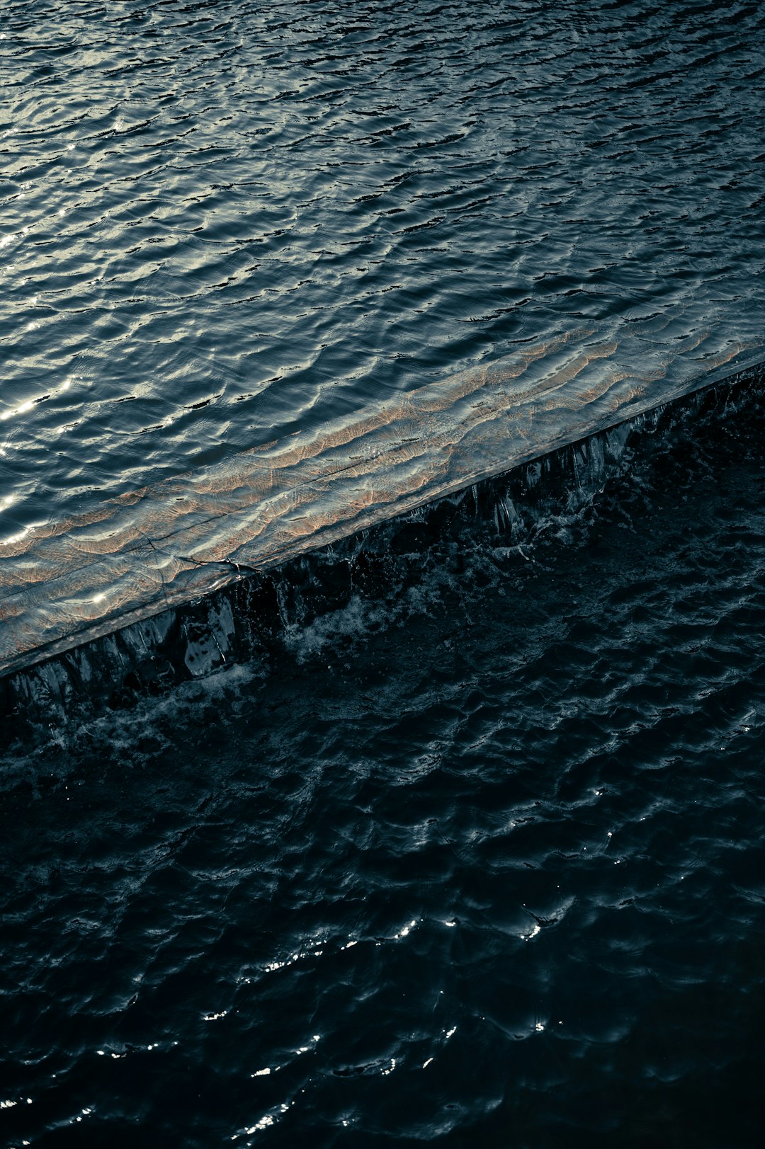 photograph of dark water surface with small waves, edge is made out of wood and metal, evening light, close up, aerial view –ar 85:128