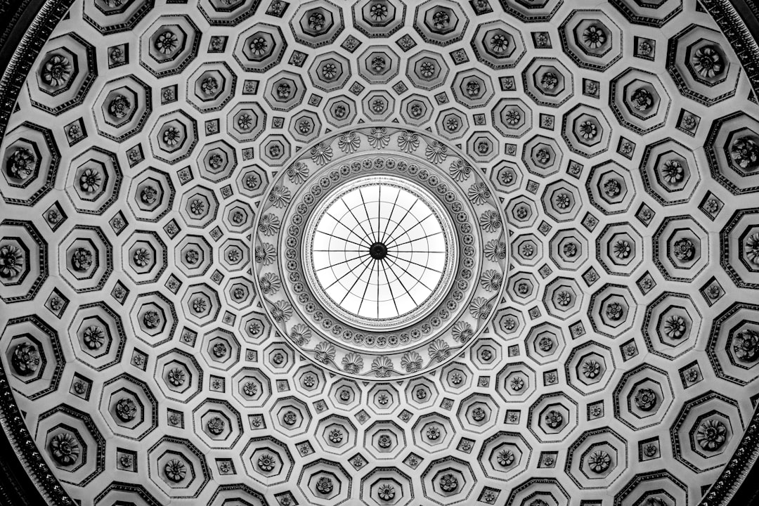 black and white photo of the ceiling dome inElyrealistic style, the architecture is a mix between classical greek and baroque styles. The patterns on it resemble hexagons with intricate designs, giving an overall symmetrical appearance. In its center stands a large circular window that allows light to filter through from above. The whole scene exudes elegance and grandeur as if looking up at something celestial or divine. –ar 128:85