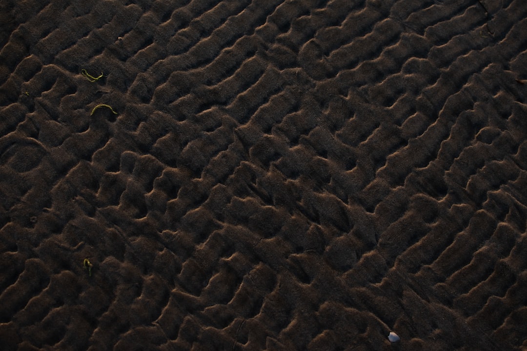 Dark brown sand ripples, top view, high resolution photograph. The sand patterns were photographed from above, showing intricate details. The ripples were styled in the manner of flowing dunes sculpted by wind and time. –ar 128:85