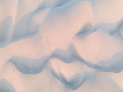 Aerial view of the surface texture of soft white sand dunes in antarctica, creating an abstract pattern with undulating shapes and shadows, captured during golden hour to enhance warm tones, shot from above by Sony Alpha A7R IV camera with G Master lens for high detail and clarity, capturing intricate textures and patterns, with soft lighting casting gentle reflections on fine particles of snow, creating a serene atmosphere, conveying tranquility and beauty of nature's artistry. --ar 4:3