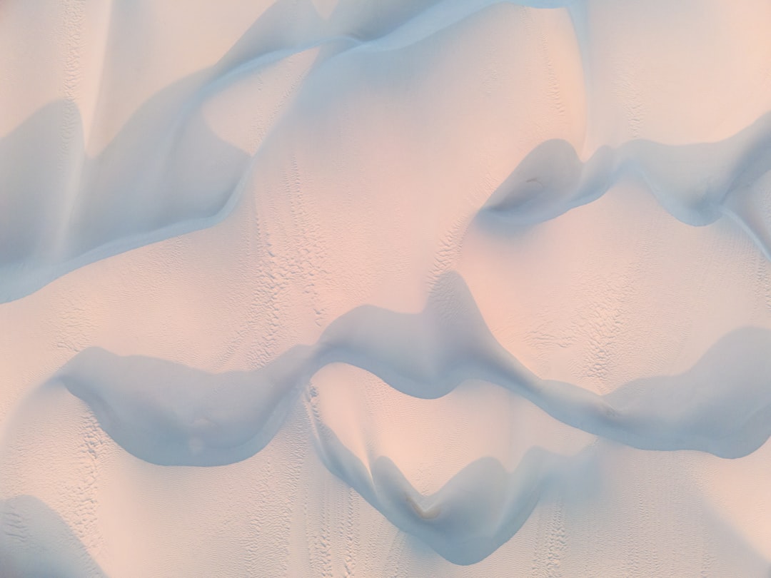 Aerial view of the surface texture of soft white sand dunes in antarctica, creating an abstract pattern with undulating shapes and shadows, captured during golden hour to enhance warm tones, shot from above by Sony Alpha A7R IV camera with G Master lens for high detail and clarity, capturing intricate textures and patterns, with soft lighting casting gentle reflections on fine particles of snow, creating a serene atmosphere, conveying tranquility and beauty of nature’s artistry. –ar 4:3