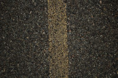 A photo of the asphalt road texture with yellow lines in the center, top view. The surface is textured and dark gray with small rocks scattered across it. There's an empty space on one side for text or design. This pattern would be suitable as background material or wallpaper to create a sense of journeying through open roads or urban streets. --ar 128:85