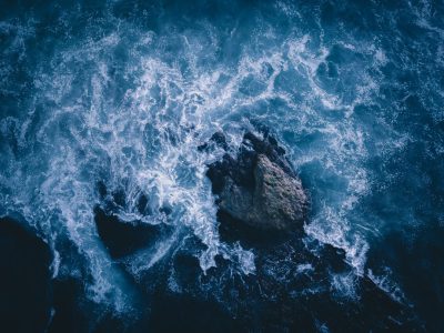 A top-down view of the ocean crashing against rocks with a dark blue and white color palette in the style of cinematic, hyper realistic photography. --ar 128:95