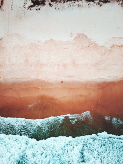 A drone photo of the beach, sand and ocean, with soft pink and orange tones, showing an area where waves can be seen crashing onto the shore, with a small silhouette visible in one corner on the right side, in a minimalistic, grainy film filter style with faded colors and a vintage style, with an ultra realistic, hyper detailed appearance. --ar 3:4