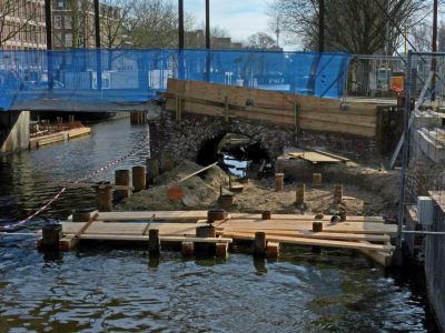 photo of construction site in Amsterdam, wooden bridge over canal being built between two underwater stone pillars with blue plastic meshing them together and around the edge is concrete on top of the old wall that used to be there before it was broken by aerial explosions during an intense urban battle. There's a small hole in one side as if someone had been shot or throw through it. In background you can see other buildings with some tiny red tape forming an tightly defined formal lines around their outside walls --ar 4:3