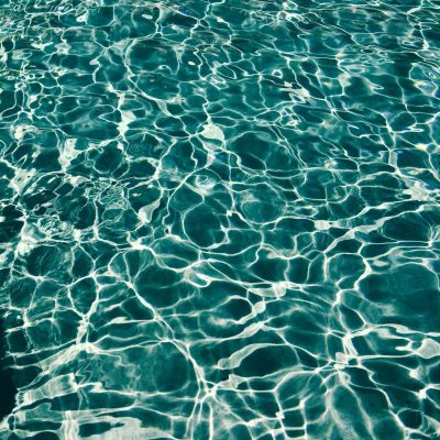 A photograph of clear teal water with ripples and reflections, taken from above. The focus is on the shimmering surface of an outdoor pool in summer. There's no one present but the light reflects off the water creating patterns that give it depth and texture. This photo was captured using a highresolution camera to capture every detail of the scene. It would be perfect for a background or wallpaper design.