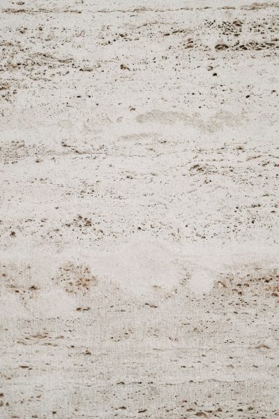 A closeup of the texture of travertine, a light grey and brown color with visible small holes and tiny cracks. The background is plain white to highlight its unique patterned surface. This top view image captures the natural beauty of the stone material. --ar 85:128